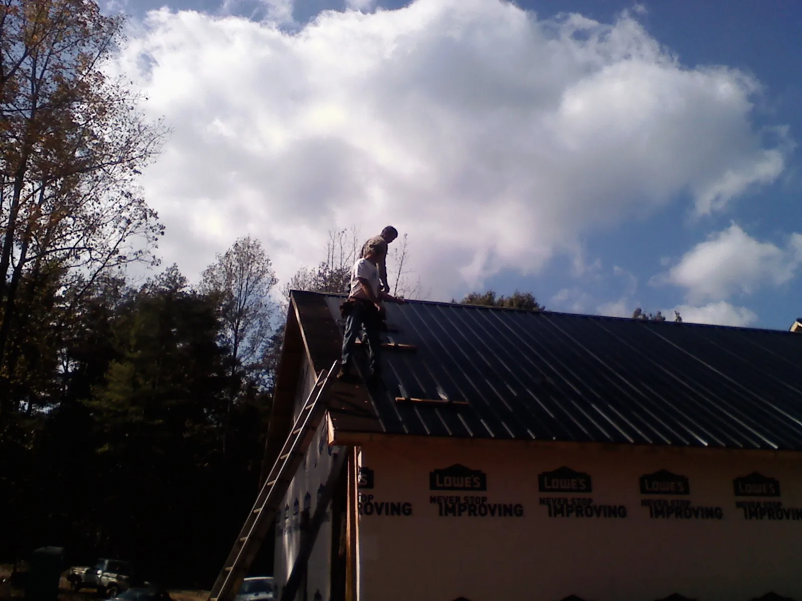 Roofer fastening dark metal panels high against a cloudy sky, from a Roof Inspection job in Slinger